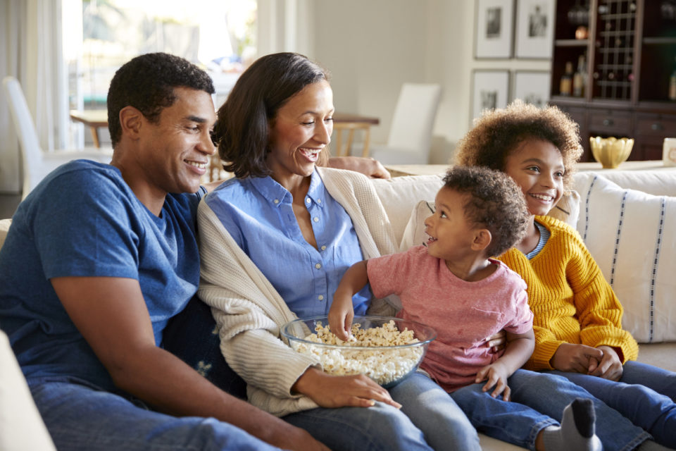 Toddler boy eating popcorn, sitting on the sofa with his sister and parents in their living room watching a movie together