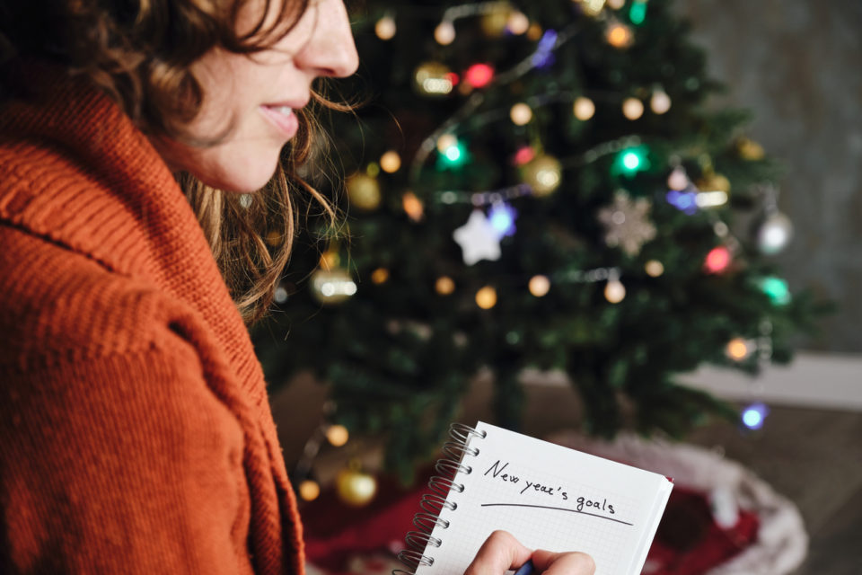 woman smiling with orange sweater in profile with new year's goals notebook in hand with unfocused christmas tree in the background