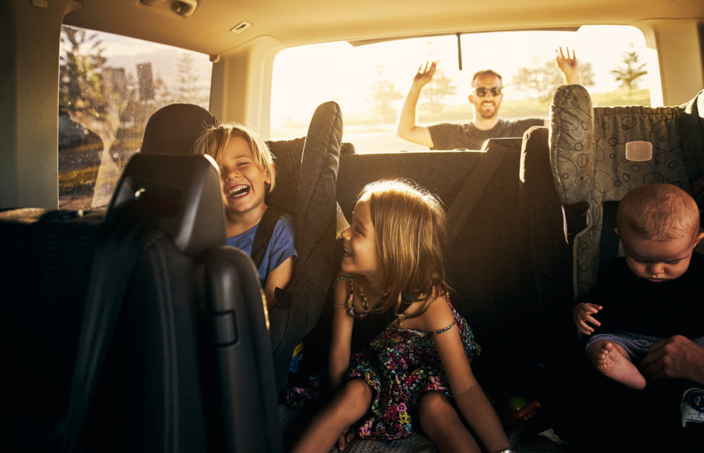 Children sitting in a car while their father packs up for a long road trip.