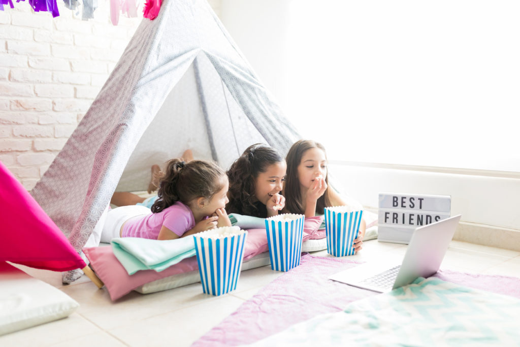 Female little friends enjoying snacks while watching film on laptop in tent during slumber party