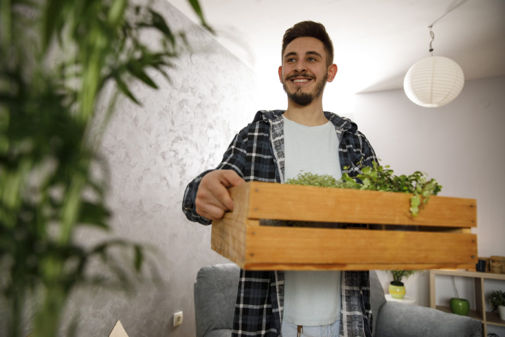 Low angle view of cheerful young man holding a wooden crate filled with potted plants and smiling while moving into his new home.